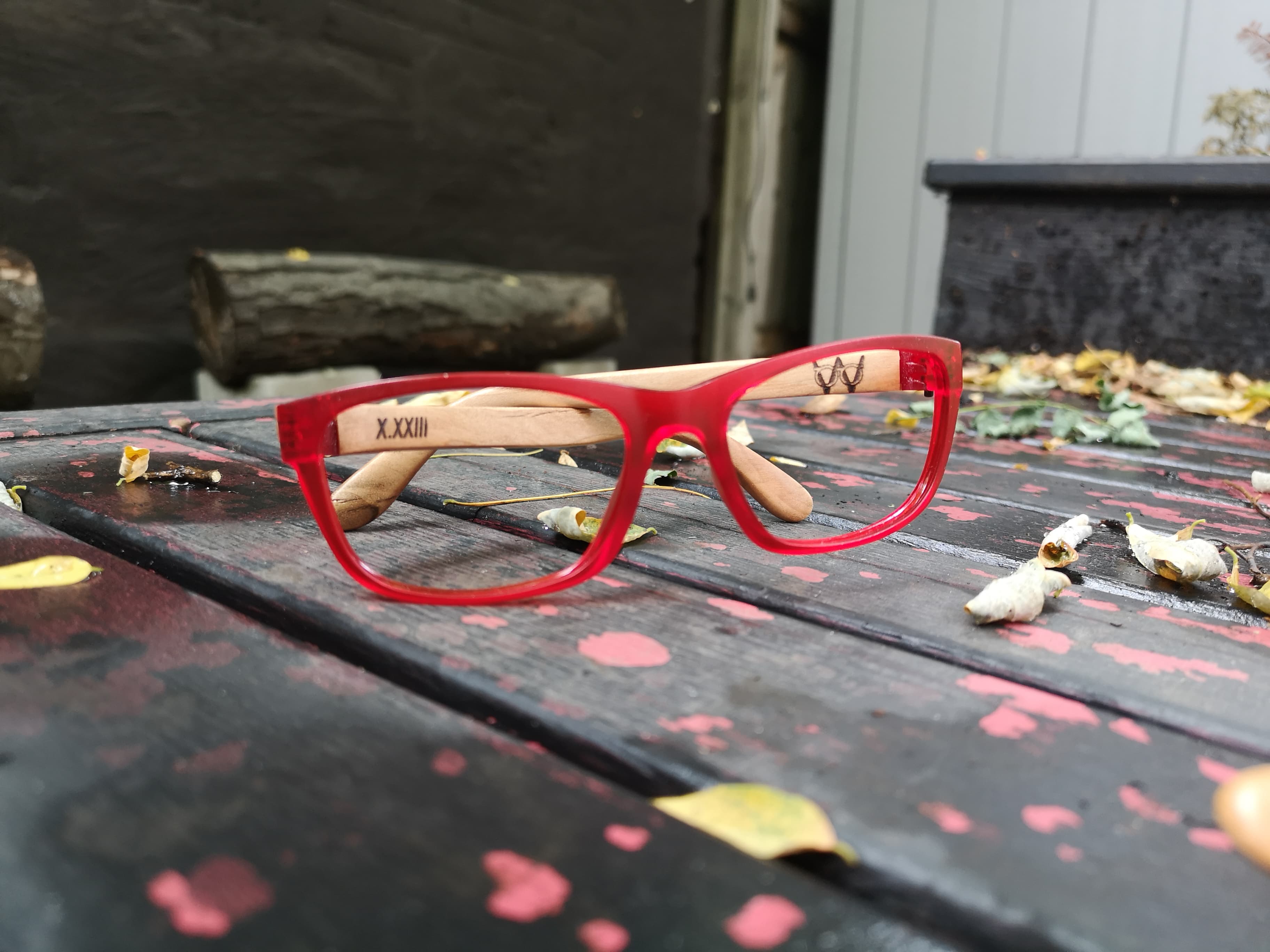 Red eyeglasses with pale maple temples engraved with a small maker's mark, photographed outdoors