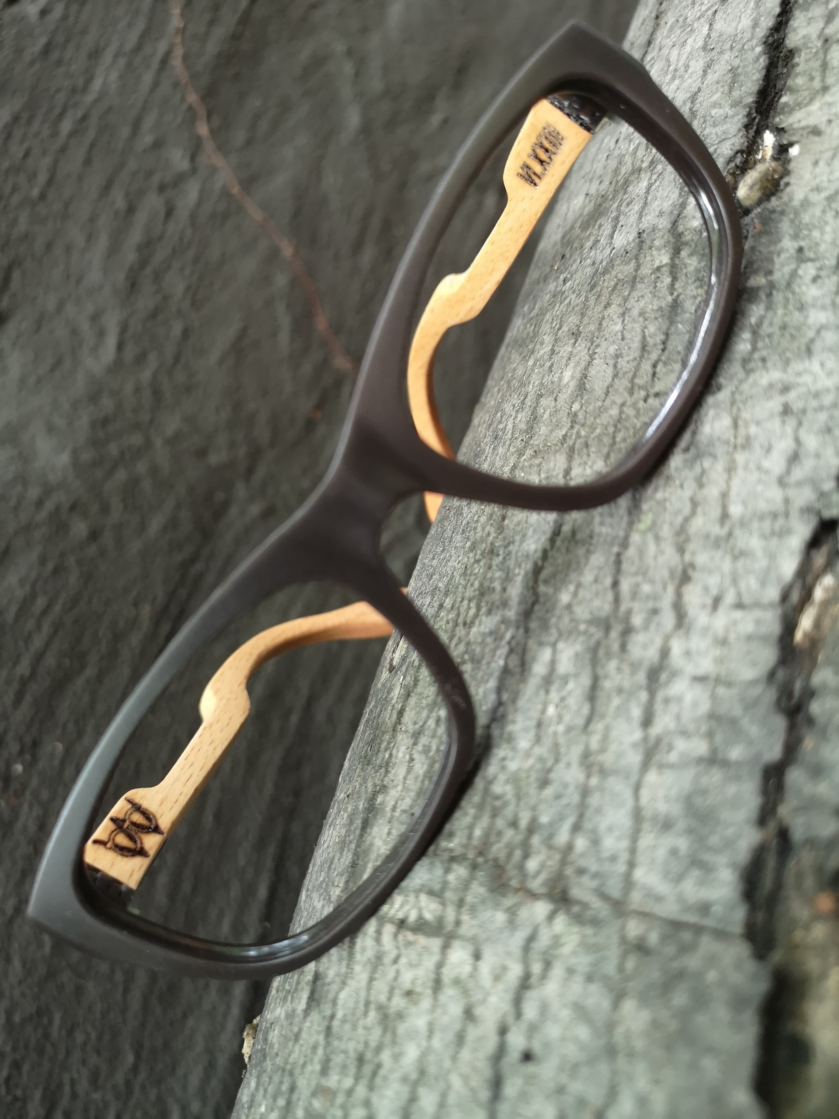 Charcoal eyeglasses with pale maple temples engraved with a maker's mark, photographed on a wood surface