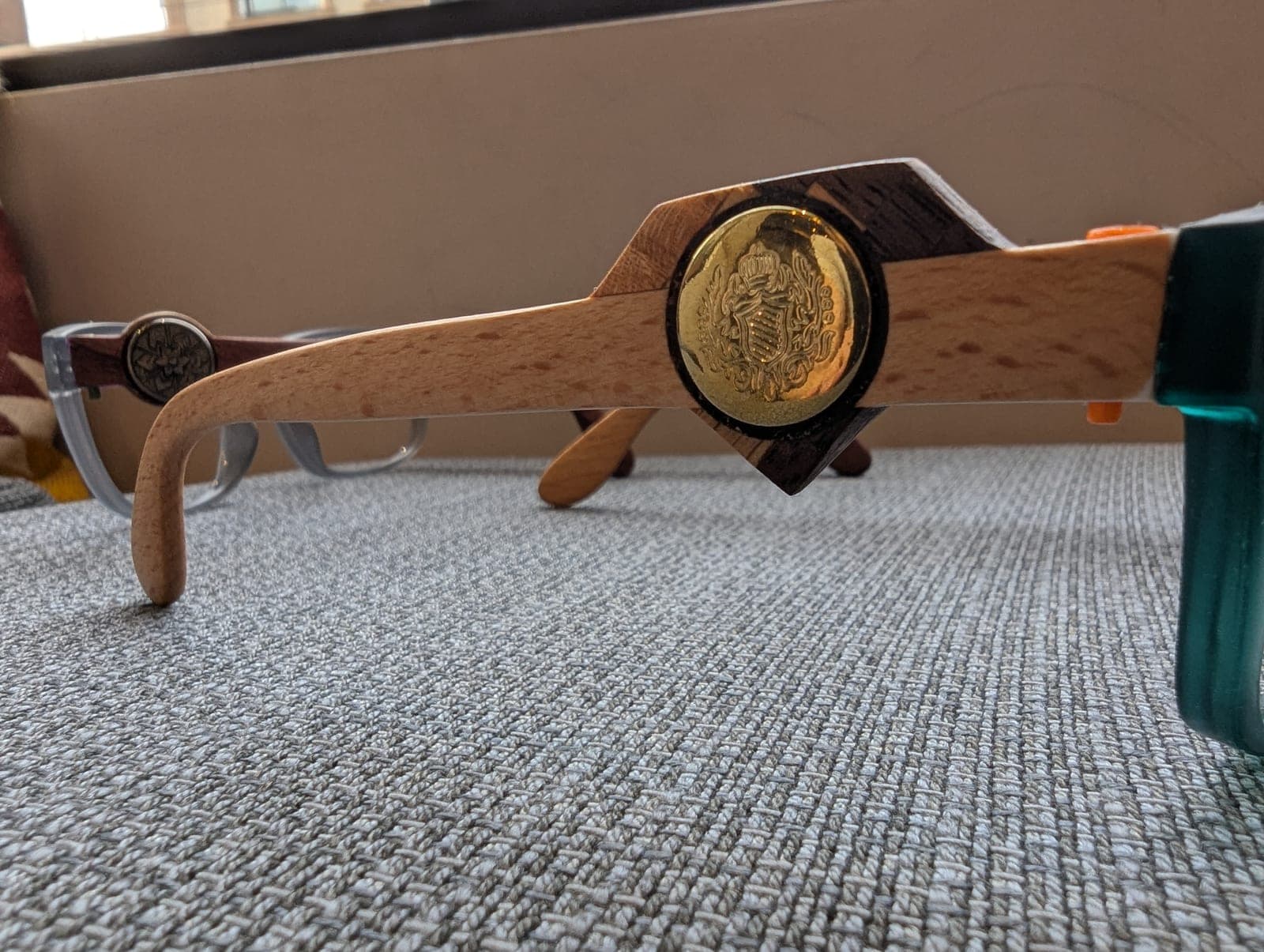Teal upcycled material eyeglass frames with light maple wood temples, each set with a round brass medallion
