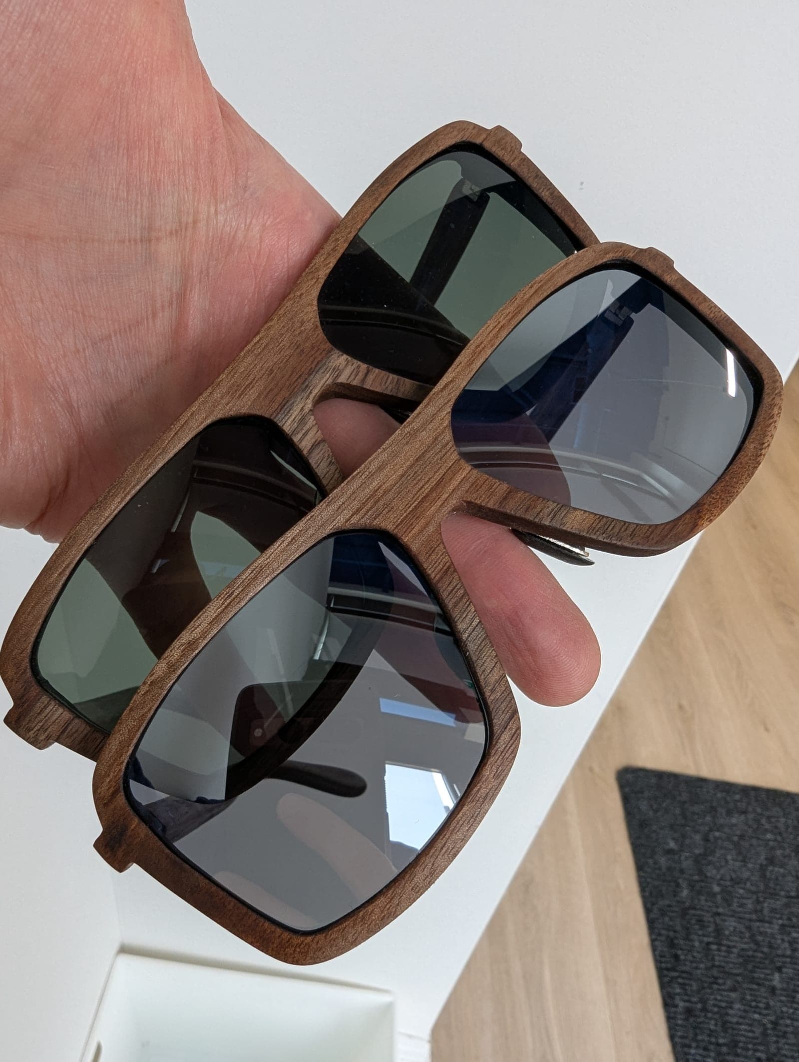 A hand holding two pairs of walnut wood sunglasses, one with green lenses and one with silver mirrored lenses